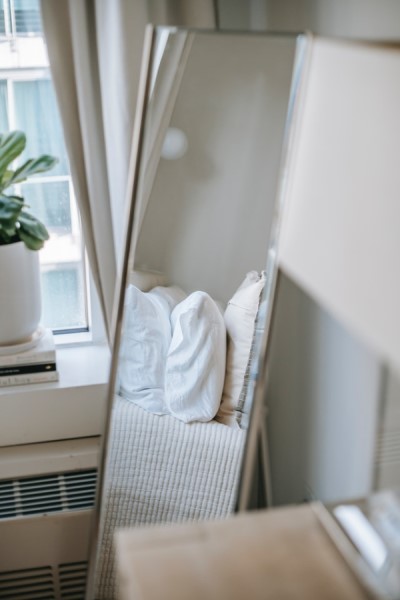 Ein minimalistischer Standspiegel in einer hellen Zimmerecke reflektiert weiße Kissen. 