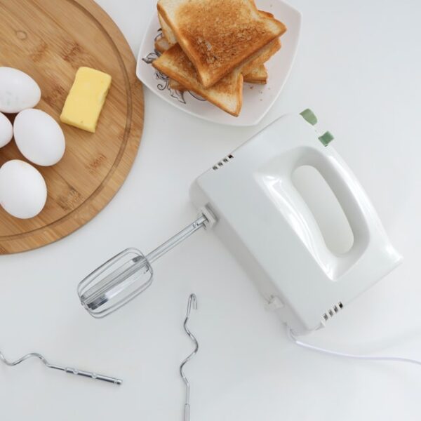Neben einem Holzbrett mit Zutaten und einer Schüssel voll Toastscheiben liegt ein weißes Handrührgerät