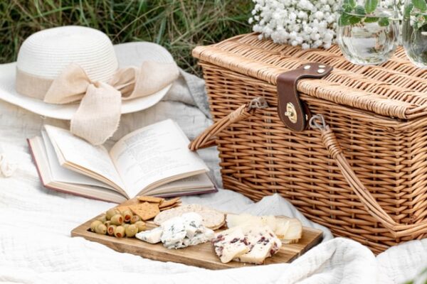 Auf einer hellen Decke liegen neben einem Picknickkorb ein Hut, ein Buch und ein paar Snacks
