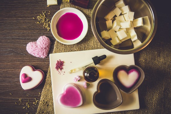 Auf einem Holztisch sind Seifen und Förmchen in Herzform zu sehen. Sowie eine Pipette, rosa Flüssigkeit und Seifenflocken.