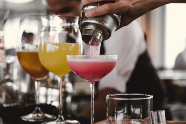 Jemand gießt einen pinken Drink aus einem Shaker in ein Cocktailglas. Im Hintergrund stehen mehr Getränke. 