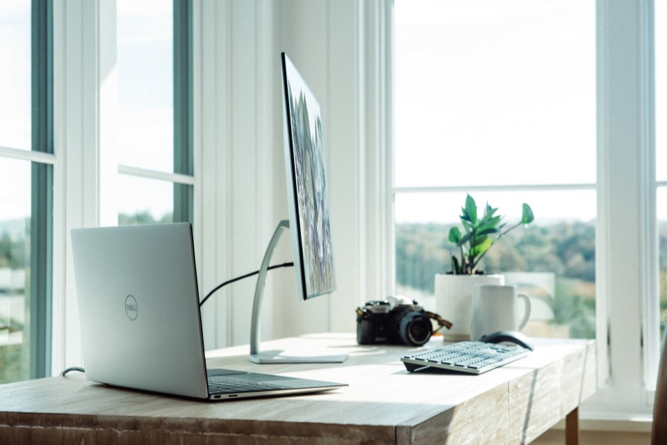 Ein ordentlicher Bürotisch mit Monitor, Laptop, Keyboard, Maus, Kamera und Zimmerpflanze. Im Hintergrund sind große Fenster.