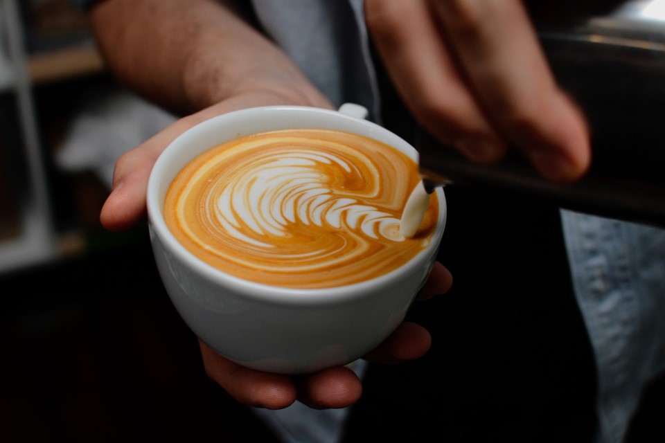 Eine Person gießt Milchschaum in eine Kaffeetasse, es entstehen helle Wellen.