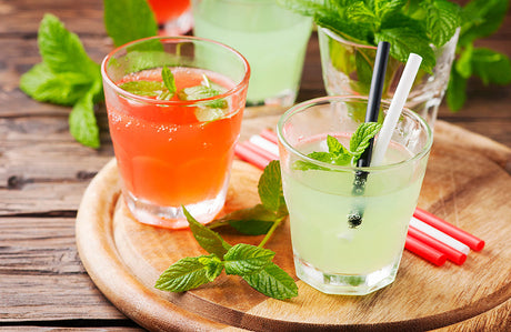 Zwei Cocktails mit Minze auf einem runden Holztablett.