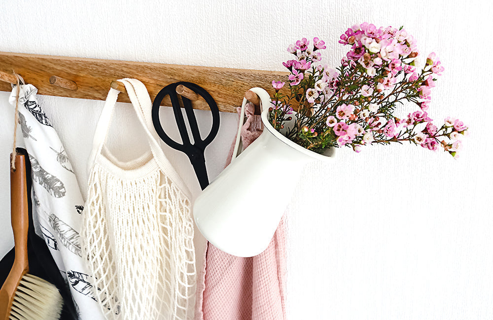 Holzleiste an einer Wand mit Holzhaken an denen Utensilien wie Einkaufsbeutel, Schere und Handtuch hängen.